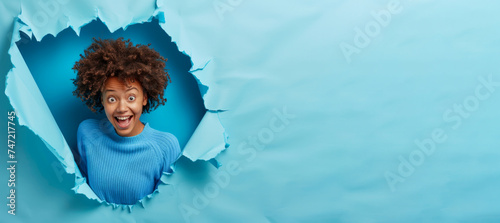 Cheerful amazed dark skinned Afro American woman indicates at blank space demonstrates advertisement wears blue jumper has happy expression breaks through paper background. Promotion concept