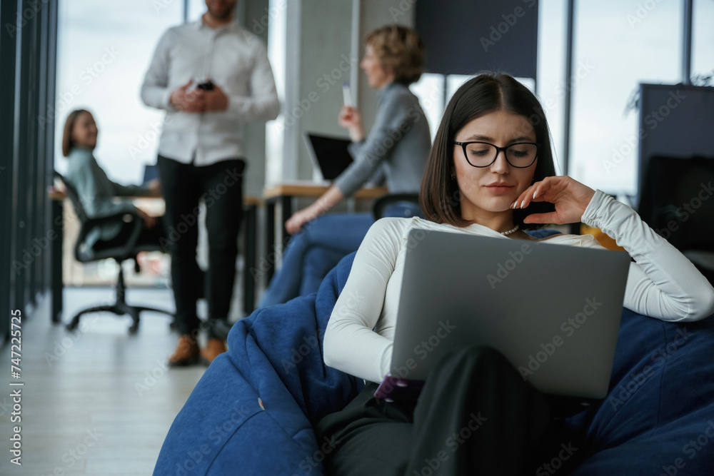 Woman in glasses is sitting with laptop. People are working in the office with bean bags chairs in it Woman in glasses is sitting with laptop. People are working in the office with bean bags chairs in it