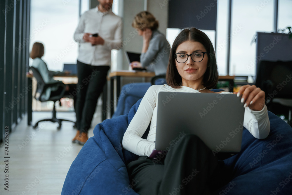 Woman in glasses is sitting with laptop. People are working in the office with bean bags chairs in it Woman in glasses is sitting with laptop. People are working in the office with bean bags chairs in it