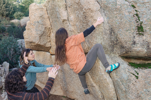 A climber reaches for a high hold, supported by attentive teammates, amidst a rugged granite boulder landscape photo