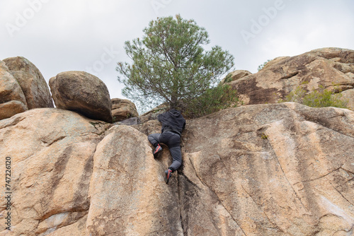 From below of anonymous climber in black attire ascends a challenging boulder, with a backdrop of rugged terrain and a solitary pine tree