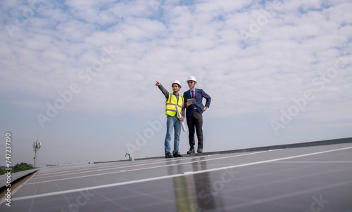 Engineers with manager walking on factory roof inspect survey and check solar cell panel by hold blueprint with turbine wind mill field background is ecology energy alternative power factory concept.