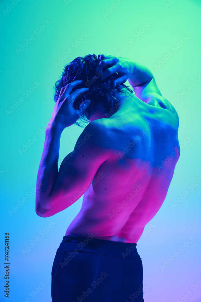 Rear view portrait of man's back in vibrant neon light against gradient blue-pink background. Model put hand on head. Concept of natural beauty people, fitness, male health and wellness, masculinity.