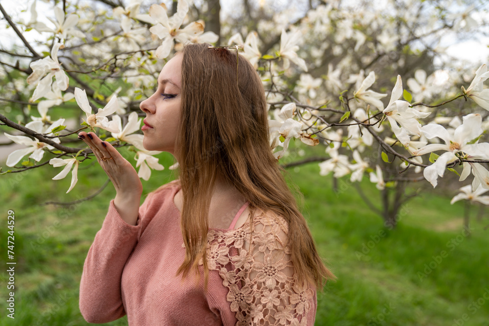 Fototapeta premium beautiful blond natural hair woman in pink outfit is posing in botanical garden park near blooming magnolia tree with flowers. Spring and purity, natural beauty. Woman Day