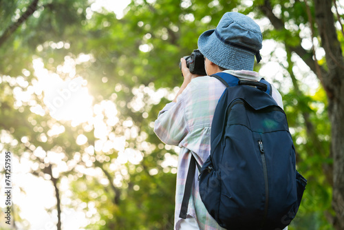 Portrait of Asian mature woman holding a camera and backpack behind her back, an Asia active senior woman enjoying nature in park. Standing on a trail in a forest outdoors. Enjoying active travel trip © Prot