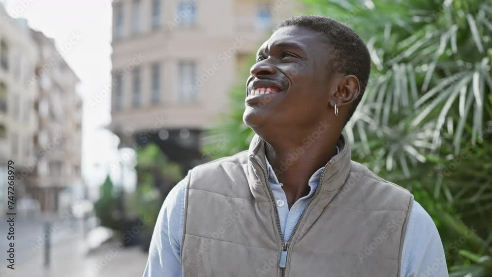 Smiling african man enjoying a sunny day outdoors in the city with green plants and a blurred urban background Smiling african man enjoying a sunny day outdoors in the city with green plants and a blurred urban background