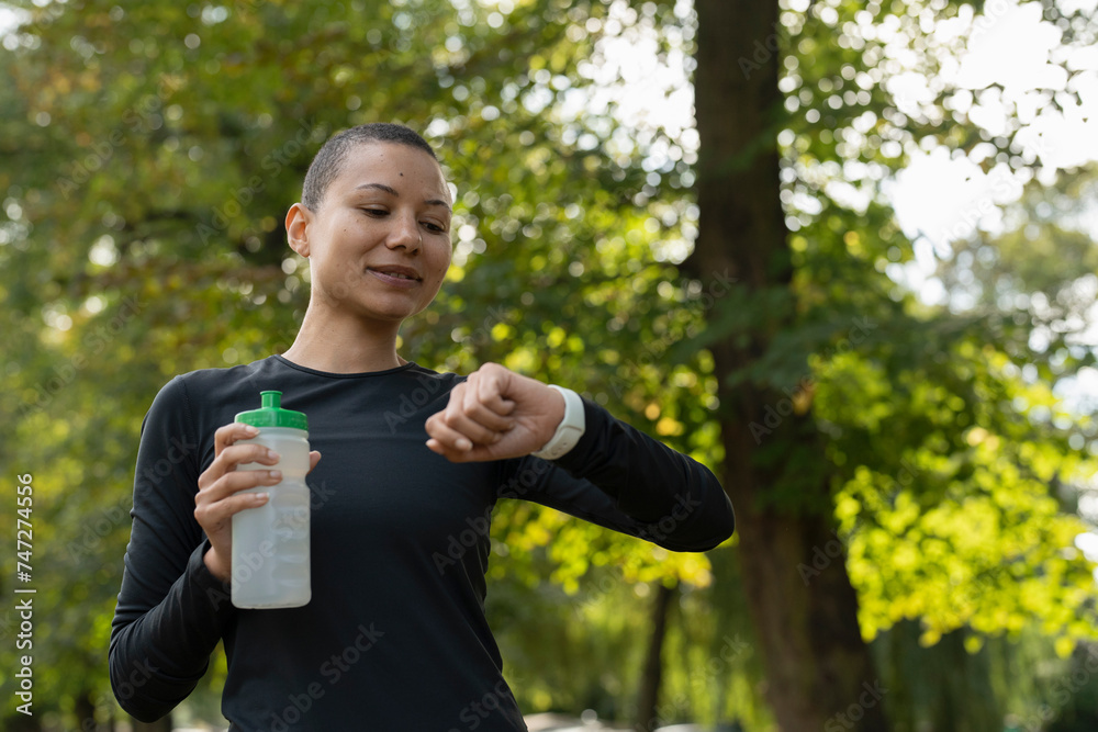 Athletic woman holding water bottle and checking smart watch in park Athletic woman holding water bottle and checking smart watch in park