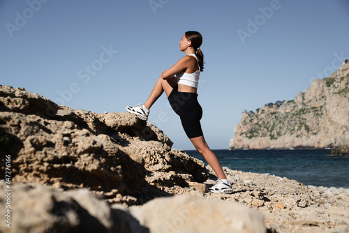 Slim fit young trainer sportswoman female athlete in fitness outfit training her legs  stretching  doing exercises for muscles outdoors by the beach shore coast. Yoga concept