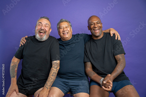 Portrait of three men laughing while sitting on bench against purple background