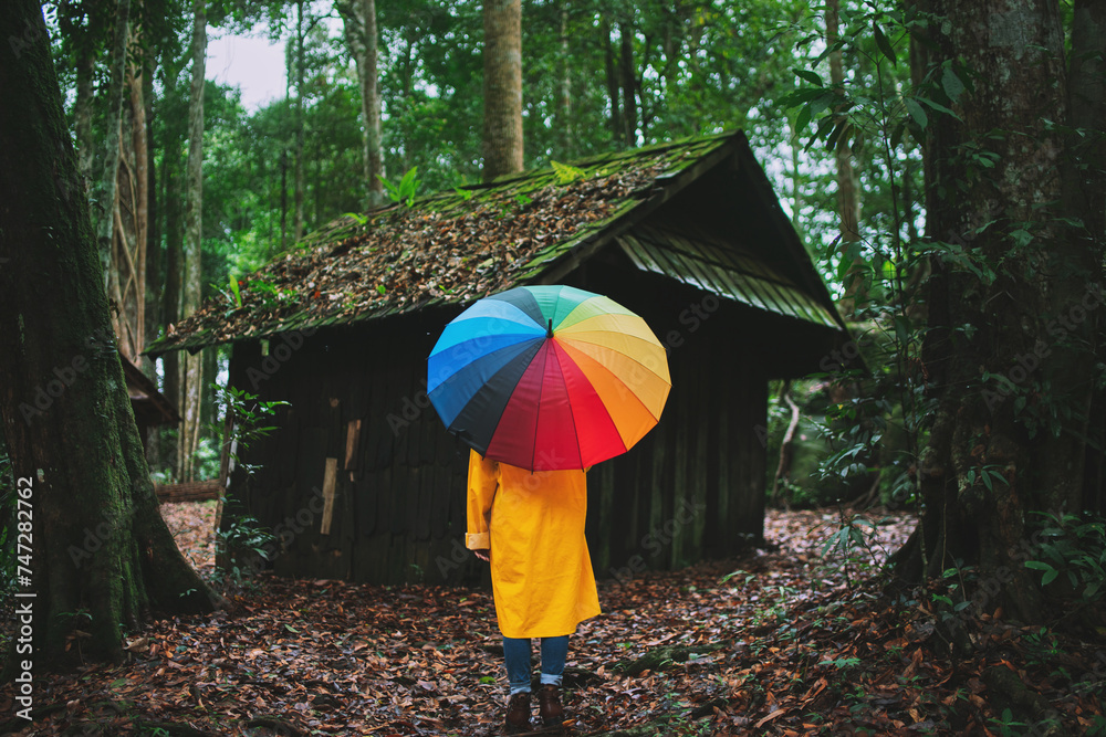 Woman holding a rainbow umbrella looking at an old hut in the forest