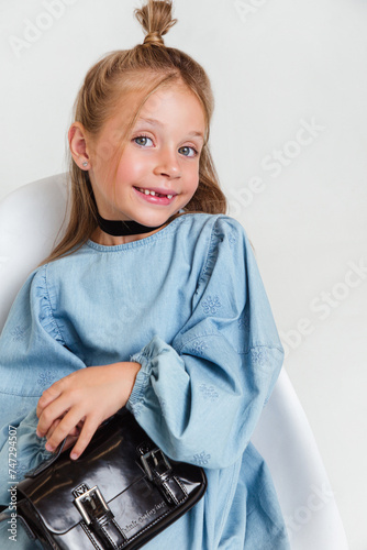 Portrait of cute toothless kid girl in blue dress with handbag on white studio background. Soft focus