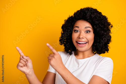Portrait of impressed girl with curly hair wear white t-shirt directing at awesome offer empty space isolated on yellow color background