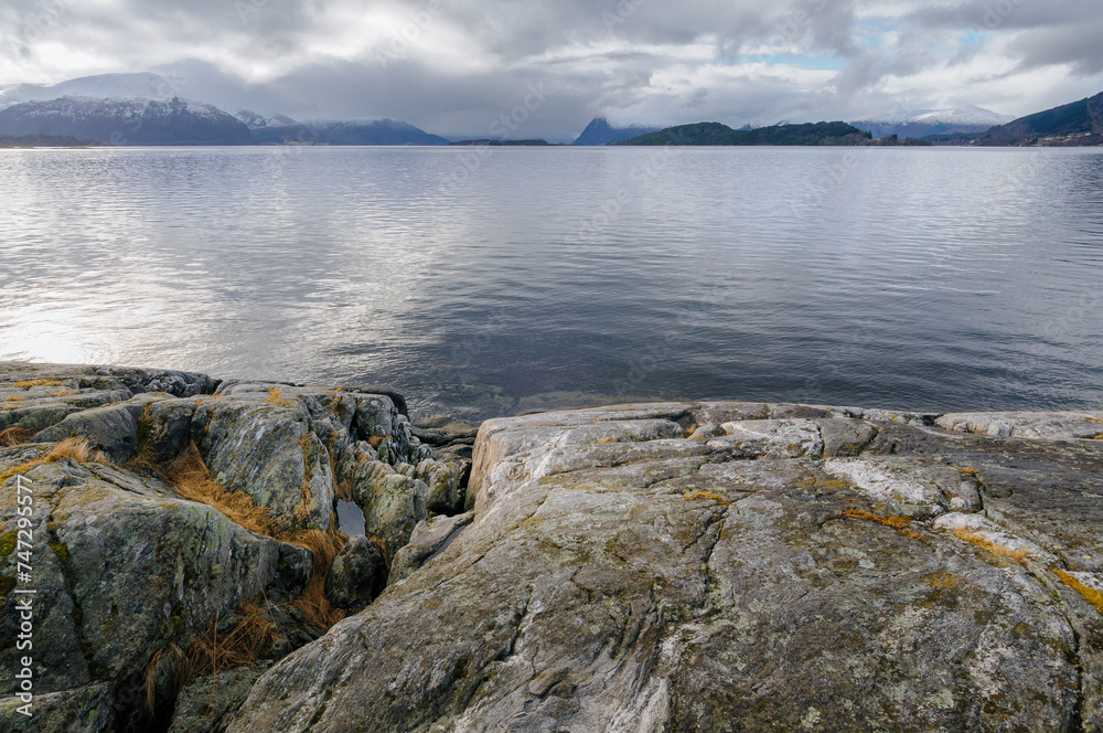 Calm water of a fjord extends to the horizon, bordered by rocky shore and distant mountains under a cloudy sky.