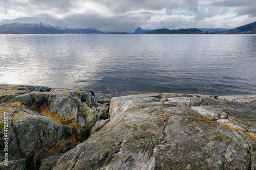 Calm water of a fjord extends to the horizon, bordered by rocky shore and distant mountains under a cloudy sky.