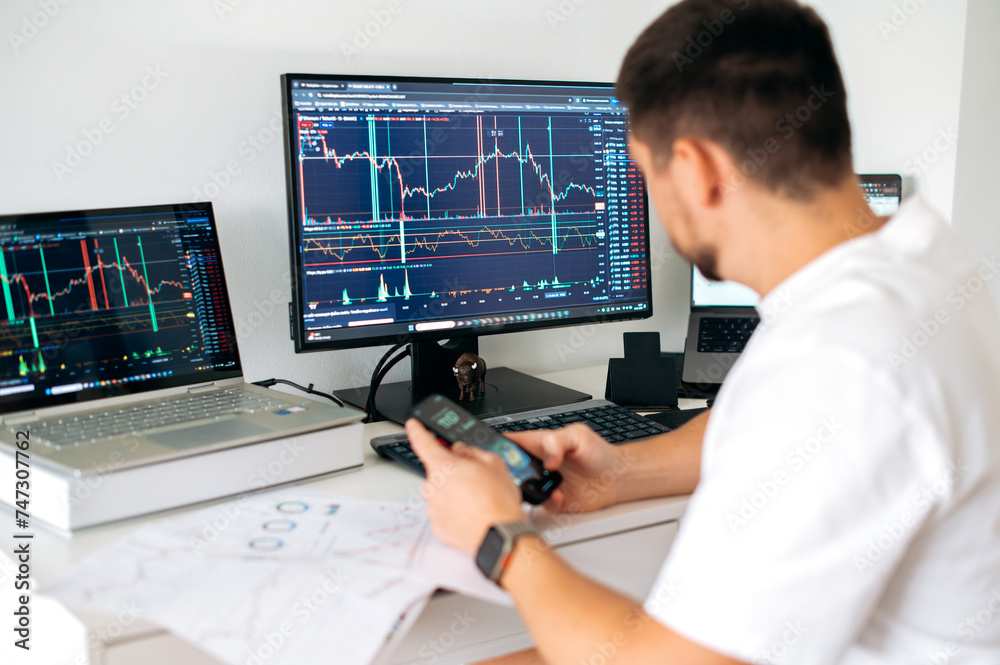 Male trader sits in home office in front of screens with graphs and financial charts, using smartphone, predicts, analyzing and studying the growth or decline of cryptocurrency.Stock exchange, trading Male trader sits in home office in front of screens with graphs and financial charts, using smartphone, predicts, analyzing and studying the growth or decline of cryptocurrency.Stock exchange, trading
