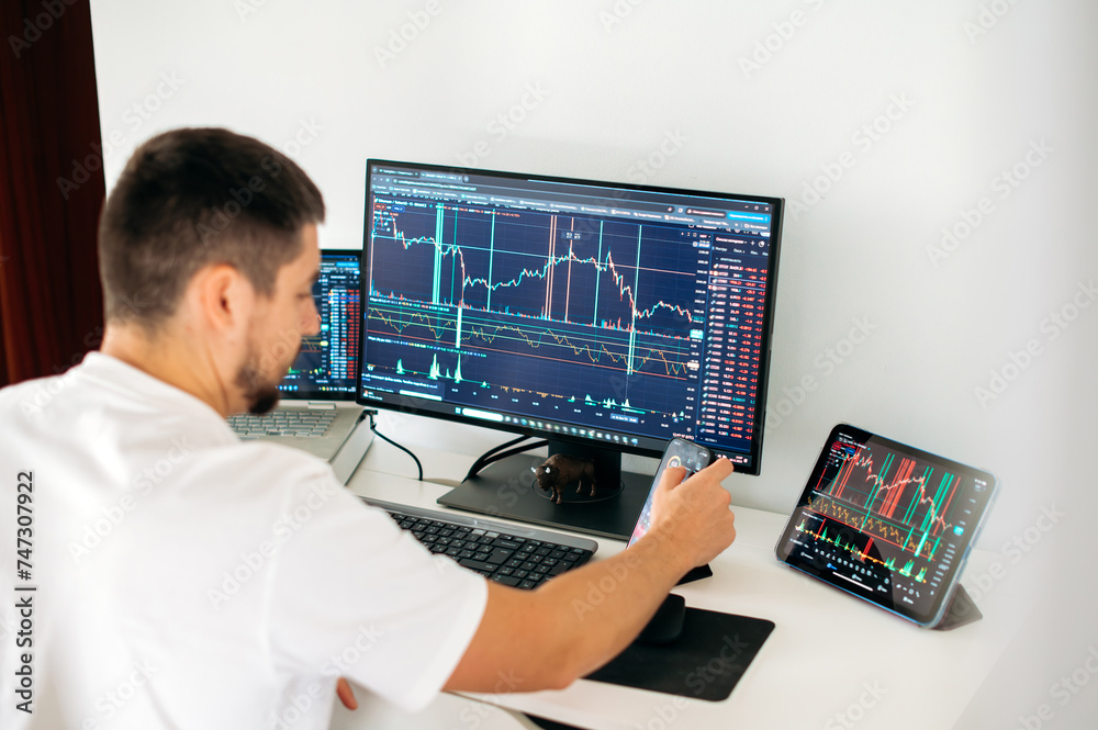 Stock trading, cryptocurrency concept. Concentrated male trader sitting at desk at home office monitoring stock market looking at monitors analyzing price flow, risks and prospects Stock trading, cryptocurrency concept. Concentrated male trader sitting at desk at home office monitoring stock market looking at monitors analyzing price flow, risks and prospects