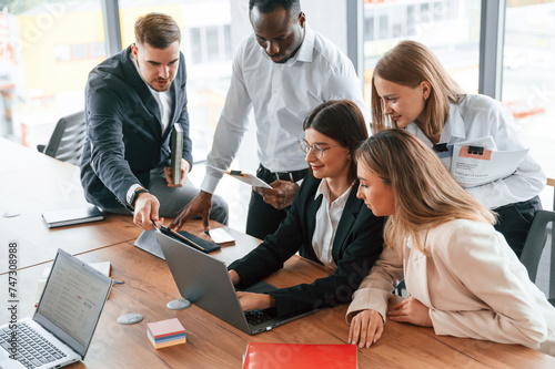 Using laptop that is on the table. Group of office workers are together indoors