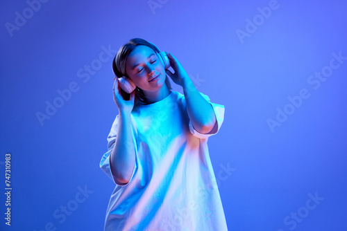 Young positive woman in casual white t-shirt listening to music in headphones against blue studio background in neon light. Concept of youth, human emotions, casual fashion