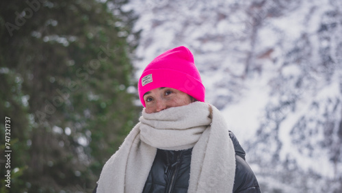 Young white woman wearing a pink winter hat and a white scarf looking to the someone else's camera while travelling to Lago di Braies in the Dolomites, Itlay, during a winter and snowy day photo