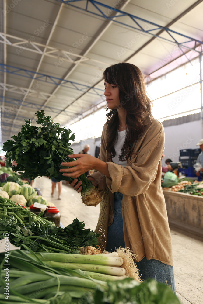 Obraz premium Young woman with reusable eco friendly net bag picking out fresh celery roots on farmers market. Conscious shopping for organic local vegetables. Close up, copy space, background.