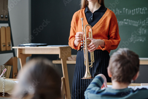 Selective focus crop shot of unrecognizable female music teacher holding brass instrument standing in front of kids in classroom