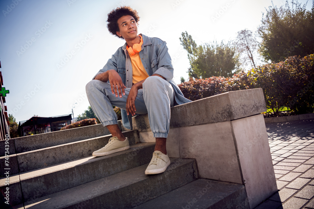 Fototapeta premium Photo of positive cheerful dreamy youngster with afro hairdo dressed jeans jacket in headphones sitting on stairs in park outdoors