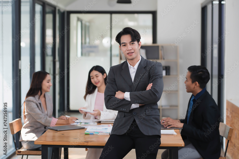 Young confident Asian businessman standing in the office room with their coworkers in the background. Young confident Asian businessman standing in the office room with their coworkers in the background.