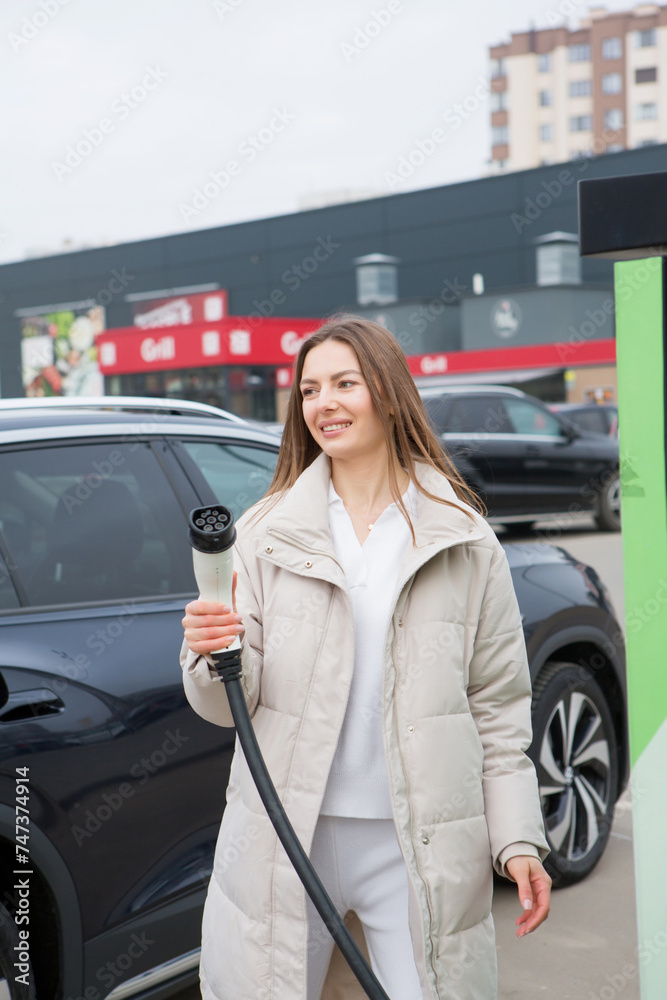 Young woman charging her electric car at the gas station, using a smartphone. Eco fuel concept. The concept of environmentally friendly transport. Recharging battery from charging station.