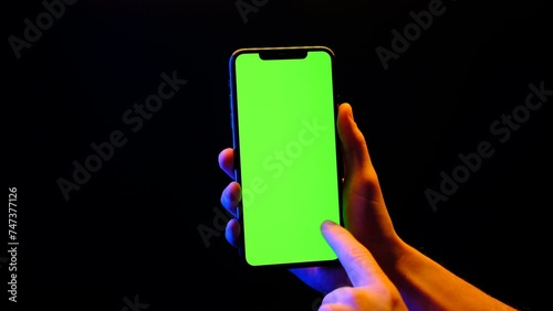 Mobile phone in hand. Holding smartphone with red blue neon lighting on dark background. Green chromakey screen with markers. Hand lifts phone up and holds it still.