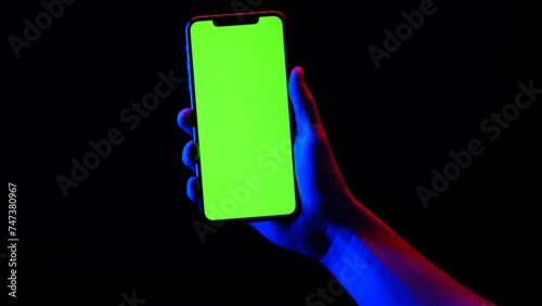 Mobile phone in hand. Holding smartphone with red blue neon lighting on dark background. Green chromakey screen with markers. Hand lifts phone up and holds it still.