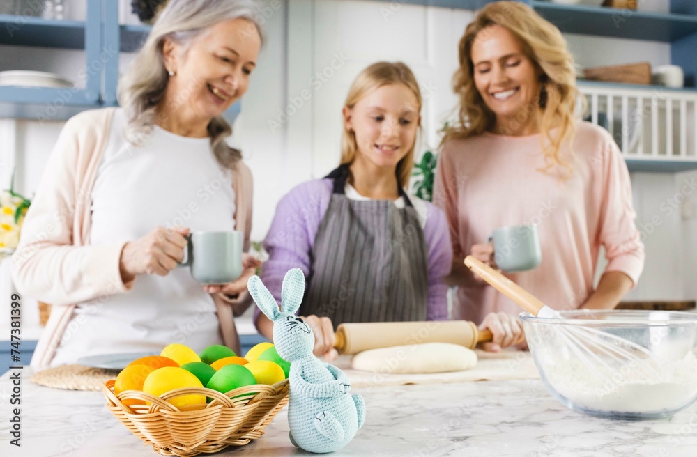 Three generations unite in kitchen, mother, grandmother, and granddaughter, as they preparing for Easter with love and laughter. Basket with painted eggs and Easter bunny in foreground Three generations unite in kitchen, mother, grandmother, and granddaughter, as they preparing for Easter with love and laughter. Basket with painted eggs and Easter bunny in foreground