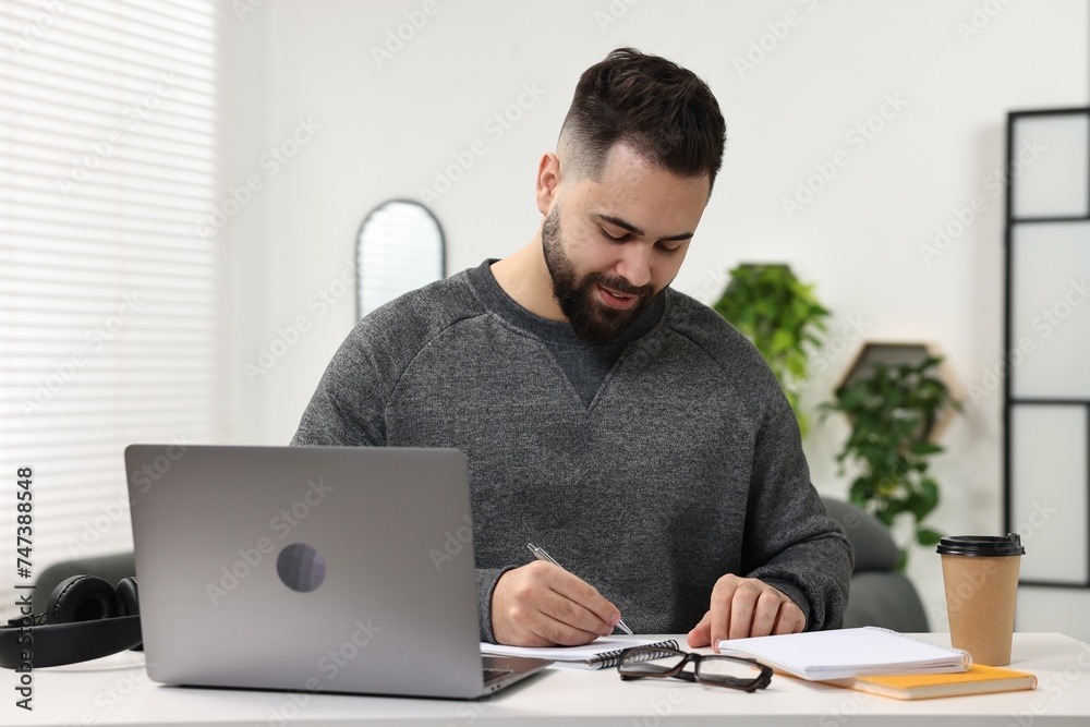 E-learning. Young man taking notes during online lesson at white table indoors E-learning. Young man taking notes during online lesson at white table indoors