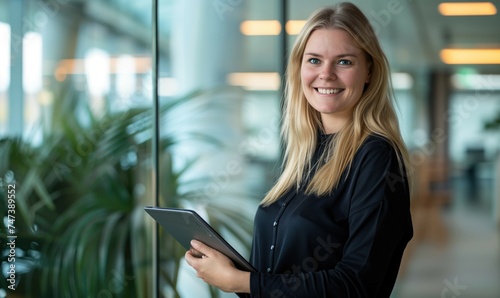 Young beautiful blond caucasian business woman or CEO executive manageer standing in light office with glass walls holding tablet and smiling at camera