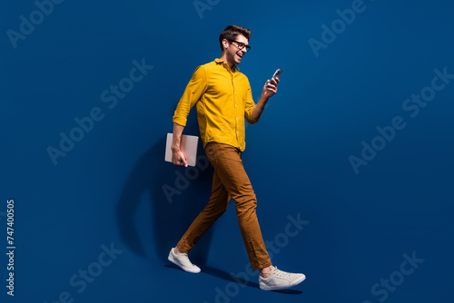 Full length photo of nice young male walk netbook hold gadget excited wear trendy yellow garment isolated on dark blue color background