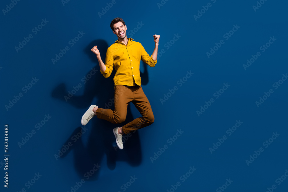 Full size photo of attractive young man jump raise fists celebrate wear trendy yellow clothes isolated on dark blue color background