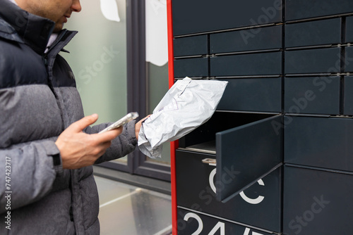 Man receiving parcel from automatic post box using smartphone outdoors. Modern delivery technologies concept. photo