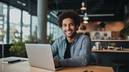 Capture the essence of a good-looking millennial office employee or student sitting at a desk in front of a laptop, smiling, and confidently looking at the camera. 