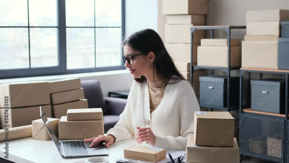 Positive Caucasian woman entrepreneur takes inventory of parcels in warehouse, enters the data in laptop. Working in office, bringing order in storage. Positive Caucasian woman entrepreneur takes inventory of parcels in warehouse, enters the data in laptop. Working in office, bringing order in storage.