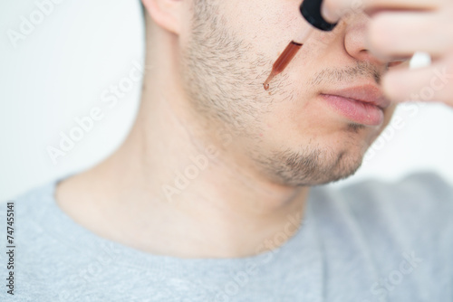 young man applying beard growth oil with pipette close up. Close-up image of handsome man holding pipette with oil for beard, Handsome man applying cosmetic serum with pipette onto face 