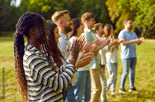 Diverse audience enjoying outdoor summer event such as music festival or community fair. Happy young Caucasian and African American people standing in park, looking away, clapping hands and smiling