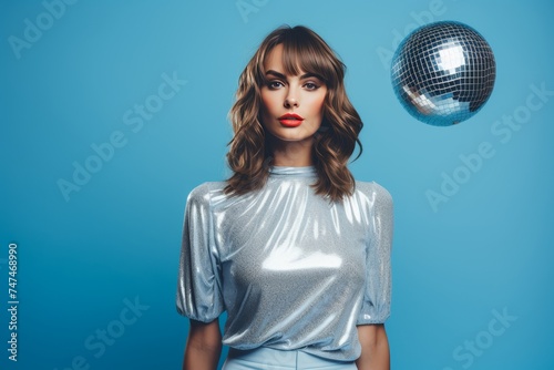 A stylish young woman in retro outfit and makeup poses confidently near a mirror ball on a blue background. She wears a silver sparkly top with balloon sleeves and high-waisted pants, exuding glamour.