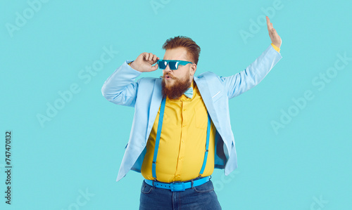 Funny chubby guy in trendy modern outfit having fun at crazy party. Happy confident fat man in cool blue spectacles, jacket, yellow shirt, and jeans with suspenders dancing on turquoise background