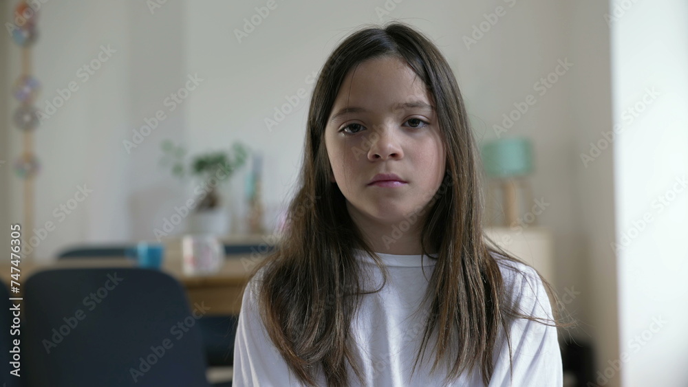 Portrait of a solemn little girl looking at camera with serious expression at home indoors, living room, 8 year old child