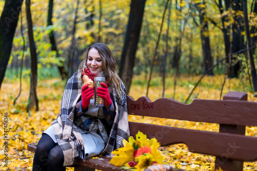A girl in an autumn park is sitting on a bench  wrapped in a blanket from the cold  drinking coffee and eating a croissant. Colorful autumn forest on the background