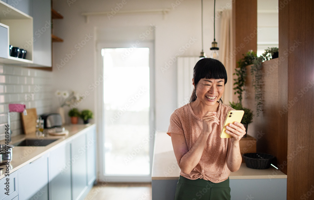 Asian woman laughing while looking at smartphone in kitchen Asian woman laughing while looking at smartphone in kitchen