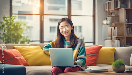 smiling chineese girl in colorful casual clothes working at a laptop at home, beige sofa, modern room space © Tetiana