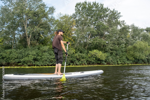 Athlete maintains graceful balance on SUP board while paddling. Man participates in aquatic workout amidst picturesque countryside