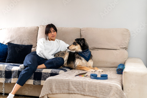 A relaxed woman sits on a sofa with her attentive dog adorned with a blue bandana, enjoying a quiet moment at home