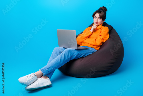 Photo of doubtful unsure woman dressed orange sweater sitting bean bag working device looking empty space isolated blue color background