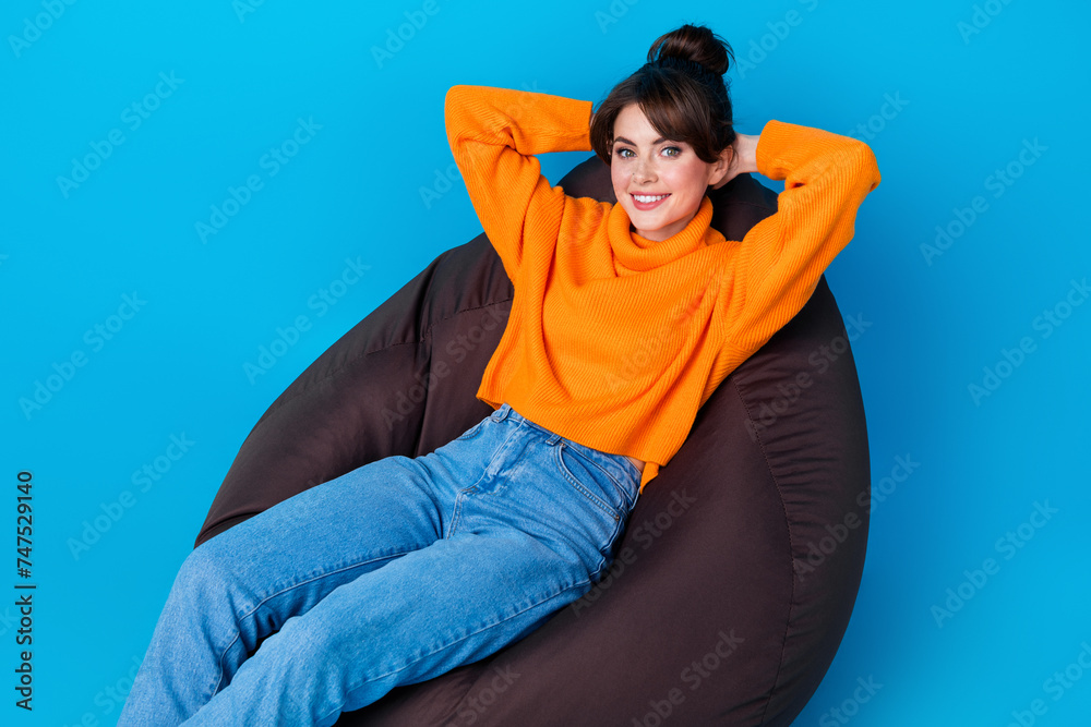 Photo of cheerful cute woman dressed orange sweater relaxing bean bag arms behind head isolated blue color background Photo of cheerful cute woman dressed orange sweater relaxing bean bag arms behind head isolated blue color background